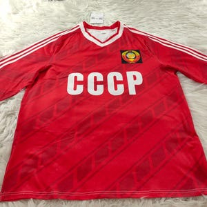 Könnte beinhalten: Ein rotes Fußballtrikot mit weißen Akzenten, das die Buchstaben "CCCP" in großen weißen Blockbuchstaben zeigt. Das Trikot hat einen V-Ausschnitt und weiße Streifen auf den Schultern. Ein Wappen ist auf der Brust zu sehen.