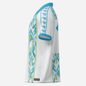 May include: White short-sleeved shirt with a turquoise and green geometric pattern. The shirt has a V-neck and turquoise trim on the collar and sleeves. A small tag is visible near the bottom hem.