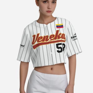 Puede incluir: Jersey de béisbol blanco con rayas negras, con la palabra "Veneka" en letras rojas y amarillas, el número "58" en negro y una pequeña bandera venezolana en el hombro derecho.