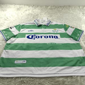 Puede incluir: Polo a rayas verdes y blancas con la palabra "Corona" en azul en el pecho. La camisa tiene cuello y mangas cortas con ribetes verdes y blancos. La camisa también tiene el logotipo del equipo en el pecho.