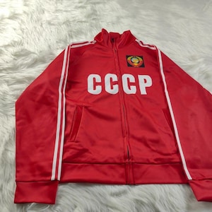 Peut inclure: Veste de survêtement rouge avec des rayures blanches sur les manches et les lettres blanches "CCCP" sur la poitrine. La veste a une fermeture éclair complète et un petit emblème sur la poitrine gauche.