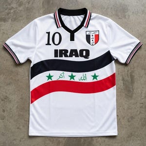 Könnte beinhalten: Weißes Fußballtrikot mit schwarzen und roten Akzenten. Das Trikot zeigt das Wort "IRAQ" in großen schwarzen Buchstaben, die Nummer "10" und ein Wappen mit arabischem Text und einem Stern. Die Ärmel und der Kragen haben rote und schwarze Verzierungen.