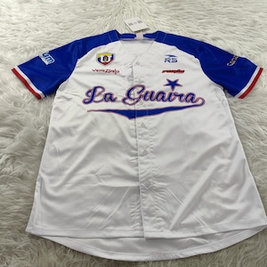 May include: White baseball jersey with blue sleeves and red trim. The jersey features the text "La Guaira" in script across the chest, along with team logos and the word "Venezuela". A tag is attached at the top.