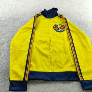 Könnte beinhalten: Gelbe Jacke mit Reißverschluss und marineblauen Akzenten sowie einem Logo auf der Brust. Die Jacke hat marineblaue Bündchen, einen Kragen und Streifen an den Ärmeln. Das Logo ist ein rundes Emblem mit den Buchstaben "CA" und einem Globus.