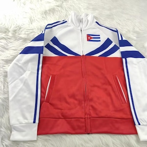 Puede incluir: Chaqueta deportiva blanca, roja y azul con cierre de cremallera completo. La chaqueta presenta un parche de la bandera cubana en el pecho y rayas azules en las mangas. Tiene dos bolsillos delanteros y un cuello alzado.