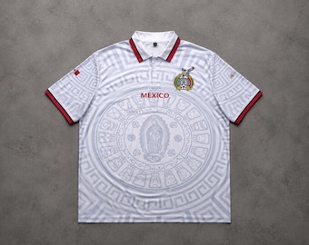 May include: A white polo shirt featuring a repeating geometric pattern, a red and black collar, and red and black sleeve cuffs. The front displays the word MEXICO in red, a central circular Aztec-style calendar design, and a crest with an eagle.
