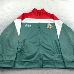 Op de afbeelding: Groene, rode en witte trainingsjas met ritssluiting, de tekst "ABA SPORT" en een voetbalembleem. De jas heeft een hoge kraag en twee voorzakken. De jas is gemaakt van een glad, glanzend materiaal.