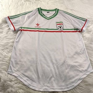 May include: White soccer jersey with green and red accents. The jersey features a V-neck collar and stripes on the sleeves. A crest with a soccer ball is on the chest, along with the brand logo. The shirt has vertical stripes.