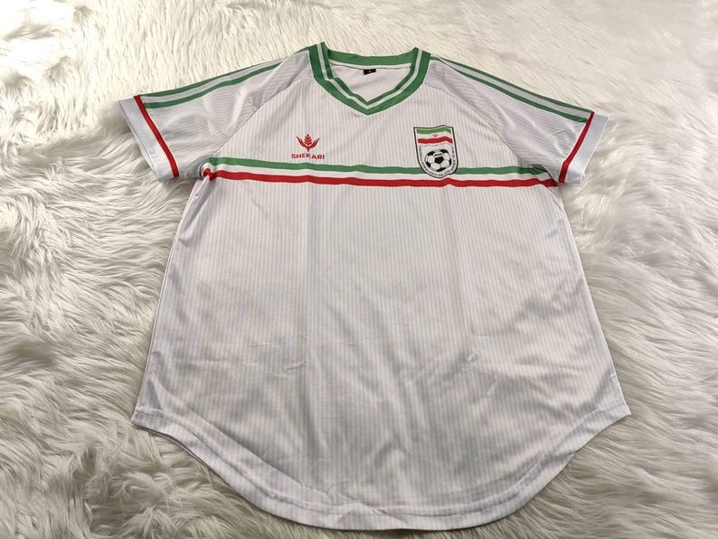 May include: White soccer jersey with green and red accents. The jersey features a V-neck collar and stripes on the sleeves. A crest with a soccer ball is on the chest, along with the brand logo. The shirt has vertical stripes.
