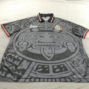 May include: Grey short-sleeved football shirt featuring a large Aztec-inspired design. The shirt has a polo collar with green, white, and red trim, matching the sleeve cuffs. The Mexican national team emblem is on the chest.
