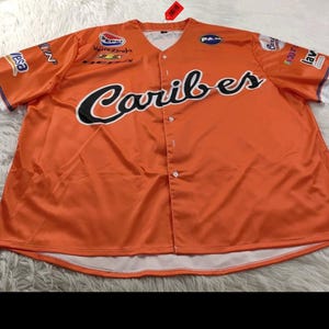 Puede incluir: Camiseta de béisbol naranja con la palabra "Caribes" en escritura negra en la parte delantera. La camiseta tiene una parte delantera abotonada, ribetes blancos en las mangas y varios logotipos. La camiseta está hecha de un material brillante.
