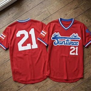 Op de afbeelding: Twee rode honkbalshirts met witte cijfers en tekst. Eén shirt heeft nummer 21 op de rug. Het andere shirt heeft het woord "Santurce" in blauw schrift en nummer 21 op de voorkant.