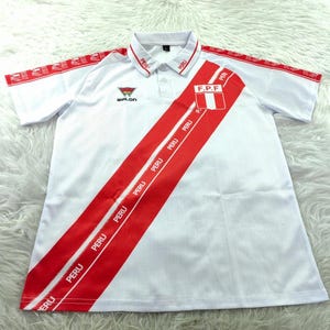 Peut inclure: Polo blanc avec des détails rouges et une bande diagonale rouge avec le mot "PERU". Le maillot a un col et des manches courtes. Le drapeau péruvien est sur la poitrine.