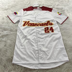 May include: White baseball jersey with red accents on the collar and sleeves. The word "Venezuela" is arched across the chest in gold lettering, with the number "24" below it in red. A small Venezuelan flag patch is on the left sleeve.