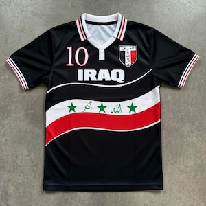 May include: Black football shirt with white and red detailing. The shirt displays the word "IRAQ" in white, the number "10" in red, and an emblem with Arabic script. The sleeves and collar have red and white stripes.