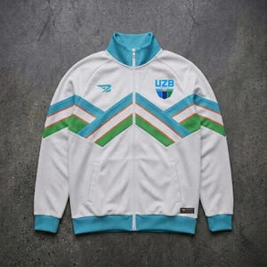Puede incluir: Chaqueta deportiva blanca con cremallera y detalles turquesa y verdes. Cuenta con cuello alto, bolsillos delanteros y las letras "UZB" con un escudo en el pecho. La chaqueta tiene un diseño retro con un patrón de chevrón.