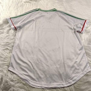 May include: White athletic jersey with green shoulder stripes and collar trim, and red sleeve trim. The jersey has a ribbed texture and a rounded hem. The shirt is displayed on a white, fluffy surface.