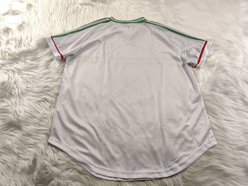 May include: White athletic jersey with green shoulder stripes and collar trim, and red sleeve trim. The jersey has a ribbed texture and a rounded hem. The shirt is displayed on a white, fluffy surface.