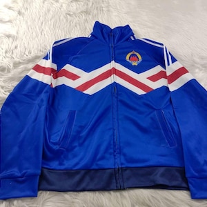 May include: A blue track jacket with a white, red, and blue chevron design across the chest. The jacket has a full zipper, two pockets, and a small emblem on the upper left chest. The jacket has a stand-up collar.