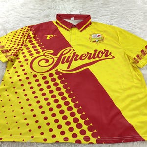 May include: Yellow and red soccer jersey with the word "Superior" in a script font. The jersey has a polo collar and a design of red polka dots on the left side. A team logo is on the right chest.