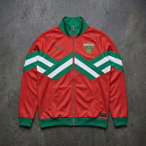 Op de afbeelding: Rood trainingsjack met groene en witte accenten. De jas heeft een volledige ritssluiting, een groene kraag en een Marokko-embleem. Het ontwerp bevat groene en witte chevronstrepen over de borst en mouwen.