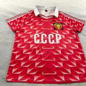 Peut inclure: Maillot de football rouge avec un col blanc et un motif géométrique. Le maillot présente les lettres "CCCP" en blanc sur la poitrine et un écusson en haut à gauche. Les manches ont des rayures blanches.