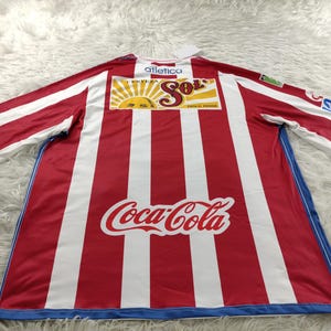 Może przedstawiać: Piłkarska koszulka w czerwono-białe pasy z niebieskimi lam&oacute;wkami. Koszulka ma duże logo Coca-Cola z przodu i mniejsze logo "Cerveza Sol" nad nim. Tekst "Atletica" jest nadrukowany nad logo "Cerveza Sol".