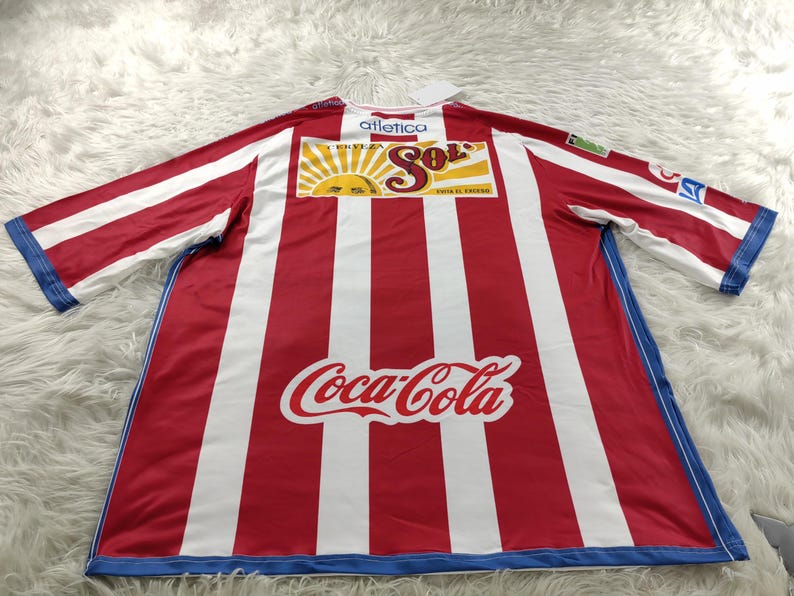 Może przedstawiać: Piłkarska koszulka w czerwono-białe pasy z niebieskimi lam&oacute;wkami. Koszulka ma duże logo Coca-Cola z przodu i mniejsze logo "Cerveza Sol" nad nim. Tekst "Atletica" jest nadrukowany nad logo "Cerveza Sol".