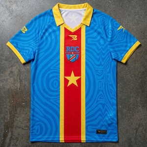 Op de afbeelding: Een blauw voetbalshirt met gele details en een rode verticale streep in het midden. Het shirt toont de letters "RDC" en een ster, met een patroon van golvende lijnen over de stof.