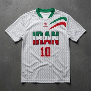 Puede incluir: Una camiseta blanca de manga corta con un patrón de chevrón gris claro. El pecho presenta la palabra IRAN en letras de bloque verdes, blancas y rojas, el número 10 en rojo, un cuello verde y un gráfico curvo rojo y verde con el texto SHEKARI.