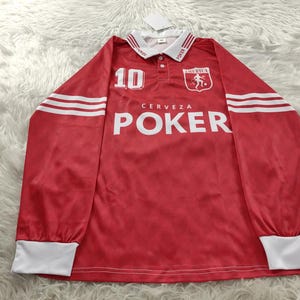 May include: Red long-sleeved jersey with white detailing. It displays the number 10, the word "POKER" in large white letters, and the text "CERVEZA" above it. The collar and cuffs are white. The jersey has an "AMERICA" crest.