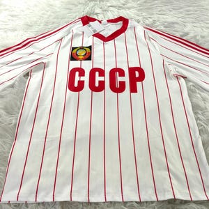 Peut inclure: Maillot à rayures blanches et rouges avec les lettres "CCCP" en rouge sur le devant. Le maillot a un col en V et une bordure rouge sur les manches et le col. Un écusson est sur le haut de la poitrine gauche.