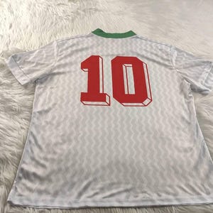 Pu&ograve; includere: Maglia bianca con colletto verde e il numero 10 in rosso sul retro. La maglia presenta un motivo sottile e ripetitivo. Il tessuto sembra leggero e adatto all'abbigliamento sportivo.