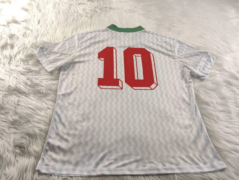 Pu&ograve; includere: Maglia bianca con colletto verde e il numero 10 in rosso sul retro. La maglia presenta un motivo sottile e ripetitivo. Il tessuto sembra leggero e adatto all'abbigliamento sportivo.