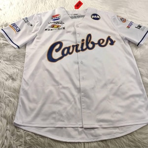 Puede incluir: Camiseta de béisbol blanca con la palabra "Caribes" en letras azul marino con ribete dorado en el pecho. La camiseta tiene varios logotipos de patrocinadores y ribetes azul marino en las mangas y el cuello. Estilo abotonado.