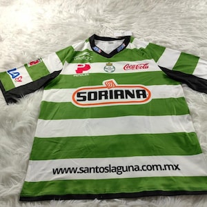 Puede incluir: Camiseta de fútbol verde y blanca con el logotipo del equipo y los patrocinadores, incluyendo "Soriana" y "Coca-Cola", y el sitio web "www.santoslaguna.com.mx".