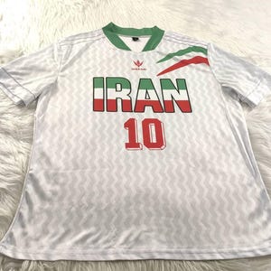 Pu&ograve; includere: Maglia da calcio bianca con colletto e finiture verdi. La parola "IRAN" &egrave; visualizzata in verde, bianco e rosso, con il numero "10" sotto in rosso. La maglia presenta un sottile motivo a zig-zag.