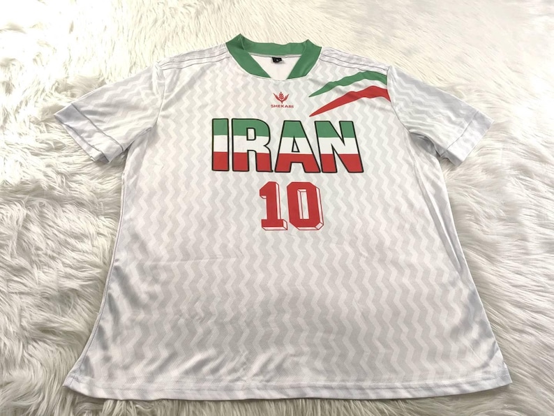 Pu&ograve; includere: Maglia da calcio bianca con colletto e finiture verdi. La parola "IRAN" &egrave; visualizzata in verde, bianco e rosso, con il numero "10" sotto in rosso. La maglia presenta un sottile motivo a zig-zag.