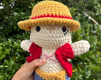 May include: A crochet plush doll featuring a cream-coloured body, a yellow straw-style hat with a red band, a red open vest, and blue shorts with a yellow sash. The doll has black bead eyes and a stitched scar detail on the face and chest.