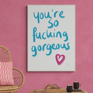 Op de afbeelding: Een ingelijste print met de tekst "you're so fucking gorgeous" in blauw schrift, met een roze hart. Een geweven stoel met een rood-wit gestreept kussen staat op een wit tapijt. Een ronde geweven tafel met zwarte mokken.