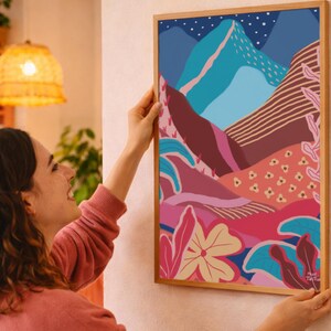 Op de afbeelding: Een ingelijste kunstdruk met een abstract landschap met bergen, bloemen en een sterrenhemel. Het kunstwerk gebruikt blauwe, roze en bruine tinten, geplaatst in een houten frame. De print wordt tegen een muur gehouden.