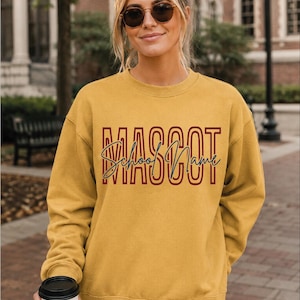 May include: A mustard yellow crewneck sweatshirt featuring the word MASCOT in large, bold, dark red block letters with a thin outline. Overlaid across the center is the text School Name written in a dark blue cursive script font.