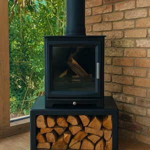 May include: A black wood-burning stove with a glass door, chimney, and a storage area for firewood. The stove is set against a brick wall and a window with a view of green foliage. The stove is on a wooden floor.