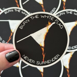 Puede incluir: Una pegatina redonda negra con el texto blanco "BURN THE WHITE FLAG" y "NEVER SURRENDER". El diseño muestra una bandera blanca en llamas. La pegatina está sujeta por una mano con esmalte de uñas negro.