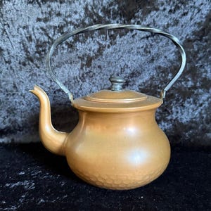 May include: A copper-coloured teapot with a hammered texture on the lower half. It features a curved spout, a lid with a small knob, and a twisted metal handle. The teapot is a warm, metallic colour.