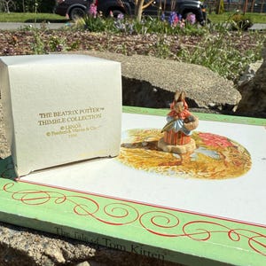 Può includere: Una scatola bianca con lettere dorate che recitano "The Beatrix Potter TM Thimble Collection Lenox Frederick Warne & Co. 1996". La scatola è appoggiata su un libro intitolato "The Tale of Tom Kitten". Una piccola statuetta di porcellana di Peter Rabbit che tiene un cestino è appoggiata sul libro.