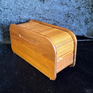 May include: A wooden bread box with a curved, roll-top lid. The box is made of light brown wood with a visible grain pattern. A small handle is cut into the side. The box sits on a dark surface.