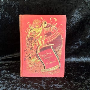 May include: A vintage book with a red cover featuring gold illustrations and text. The cover includes an illustration of a cherub and the title "Sans-Souci Illustrations de B. Monvel" by Adriana Piazza.