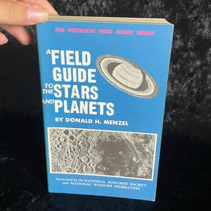 Puede incluir: Un libro de bolsillo vintage titulado "A Field Guide to the Stars and Planets" de Donald H. Menzel. La portada es azul con ilustraciones de Saturno y la luna. Patrocinado por la National Audubon Society y la National Wildlife Federation.
