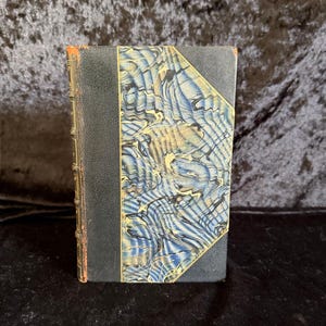 May include: A vintage book with a marbled cover in shades of blue, cream, and gold. The spine is dark blue with gold detailing. The book's edges show signs of age, adding to its antique charm.